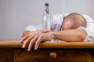 Drunk bald man wearing white shirt slouched on wooden table-empty bottle